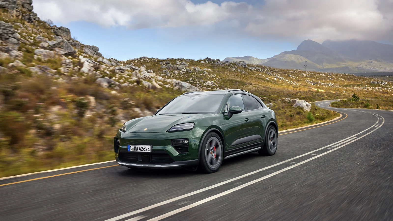 Mörkgrön eldriven Porsche Macan 4S i fart på en kurvig landsväg i ett bergslandskap.